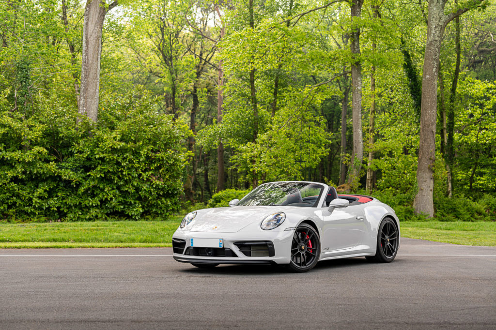 White Porsche 911 Cabriolet with its top down at the edge of a green forest — same model shown for reference